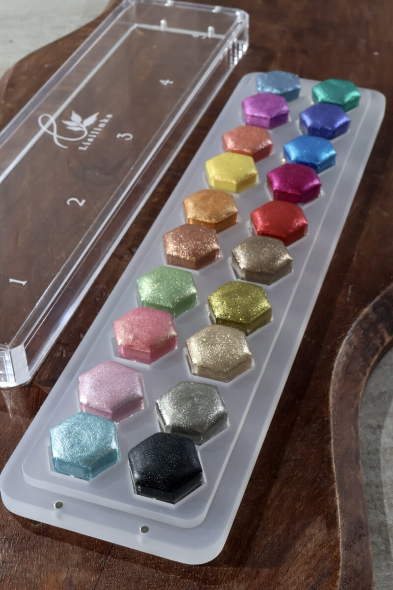 Twenty sparkling metallic watercolors in Lisilinka hexagon pans, arranged in the five palettes of Tutorial Set Vol. 2, next to the acrylic lid with the tutorial numbers 1 to 5 engraved